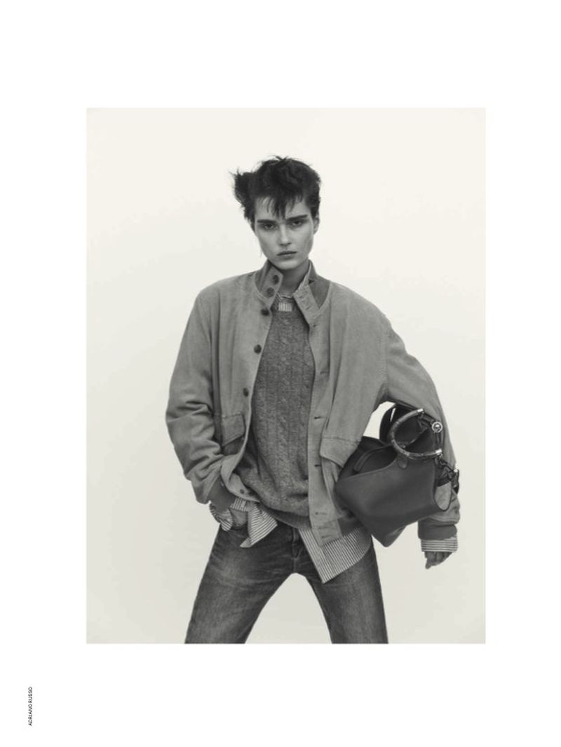 Model wearing a button-down jacket and jeans holding a handbag against a plain background