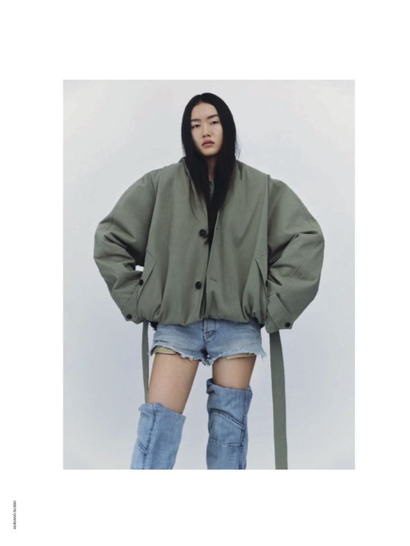 Adult female poses in an olive jacket, denim shorts, and denim boots against a white background in a studio-style fashion shot.