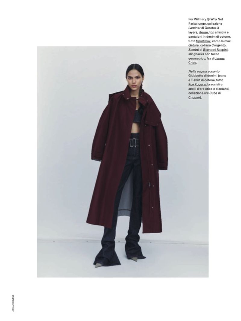 Model wearing a long burgundy coat and dark denim jeans standing against a light grey studio background.