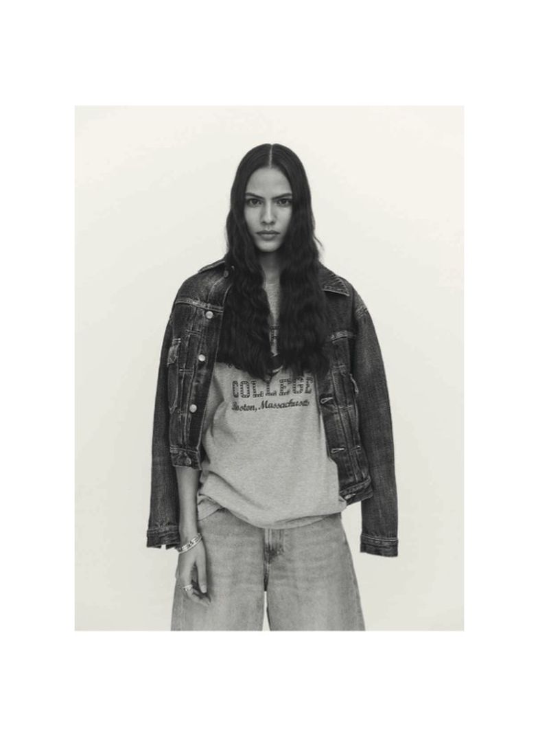 Portrait of a stylish adult female with long hair, wearing a denim jacket and college sweatshirt, posing in front of a neutral backdrop.