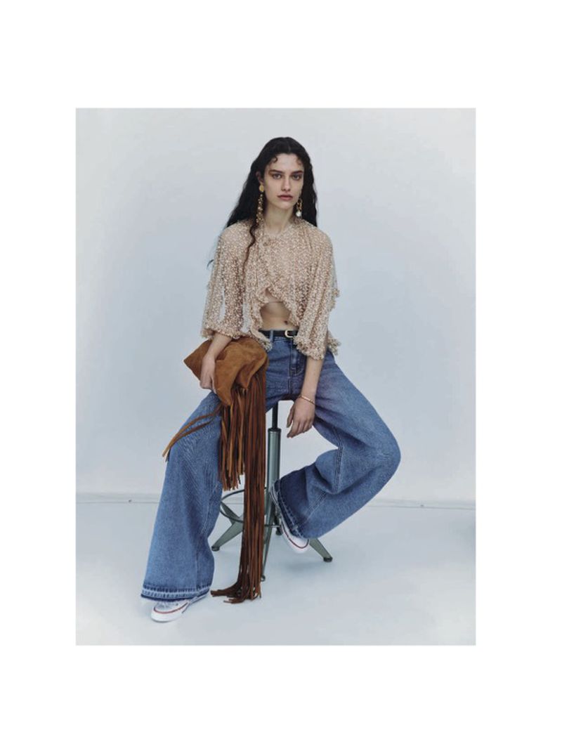 A woman sits in a photo studio wearing a lace blouse, wide-leg jeans, and sneakers while holding a fringed purse.
