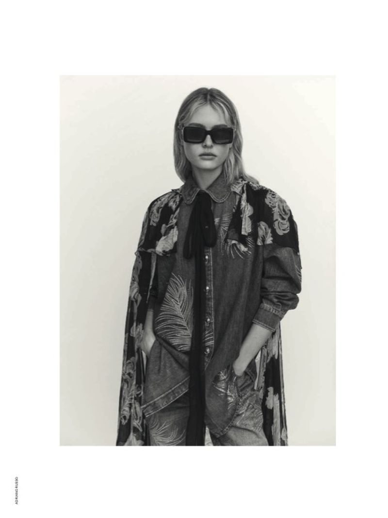 Model in dark sunglasses wearing a patterned denim shirt and long draped jacket against a neutral studio backdrop.