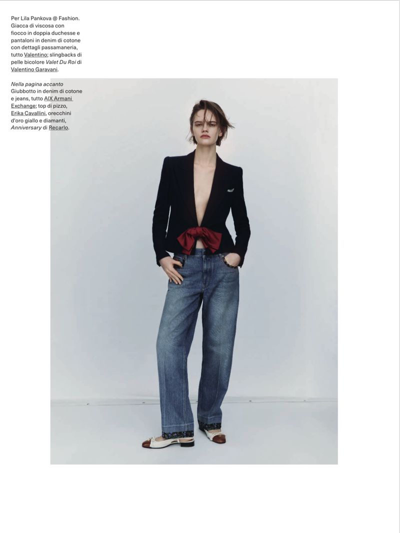 Fashion editorial shot of a woman in a dark blazer with a large bow, wide-leg jeans, and ballet flats, posing indoors against a neutral backdrop.