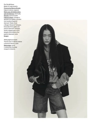 A young Asian woman poses in a fashion editorial shot, wearing a blazer, hoodie, and jean shorts, holding a black purse indoors.