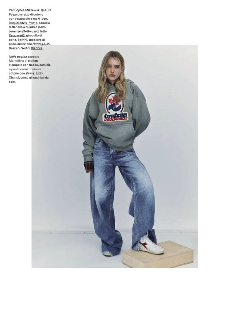A woman poses in a gray hoodie, baggy jeans, and sneakers. She is standing on a wood block against a white wall background.
