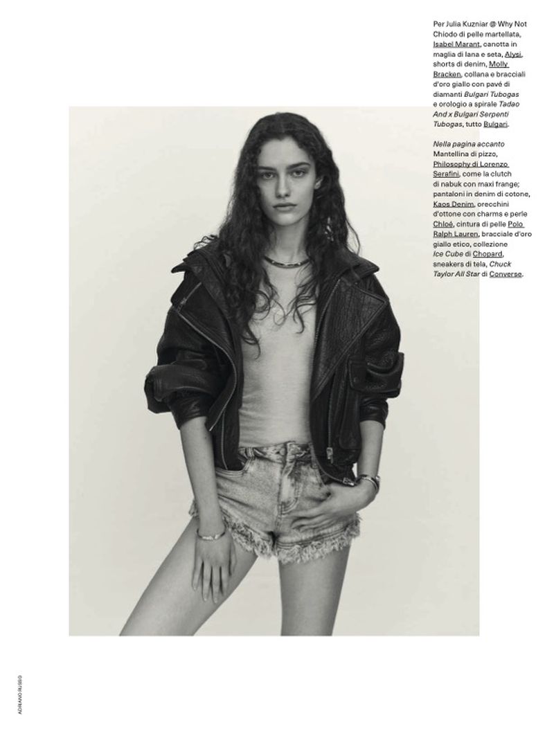 Model with long dark wavy hair wearing a black leather jacket and denim shorts against a plain background.