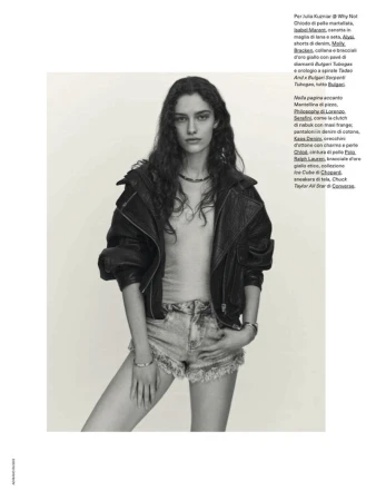 Model with long dark wavy hair wearing a black leather jacket and denim shorts against a plain background.