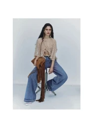 Model seated on a stool wearing a sheer beaded top, blue jeans, and holding a brown fringed suede bag against a plain grey backdrop.