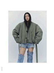 Model wearing an oversized olive green jacket, denim shorts, and tall boots against a plain backdrop.