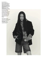 Model with long dark hair wearing a dark blazer over a grey hoodie and denim shorts, holding a black handbag.