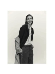 Smiling model wearing a dark blazer, white top, and grey trousers against a plain white background
