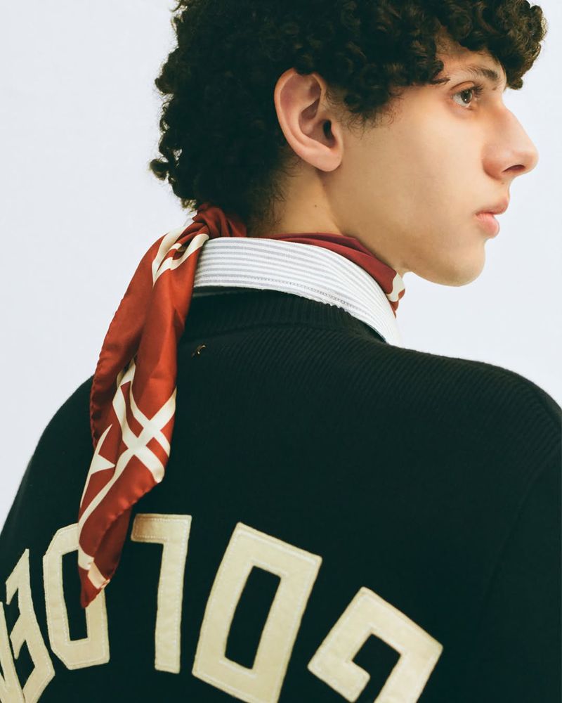 Young man in black sweater with 'GOLDEN' text poses in a white photo studio, styled with a burgundy scarf and striped collared shirt.