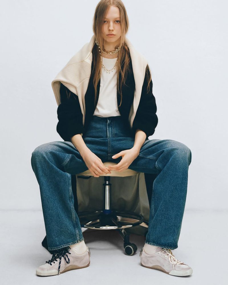Model seated on an office chair wearing wide-leg blue jeans, a black jacket, and layered necklaces against a white studio background.