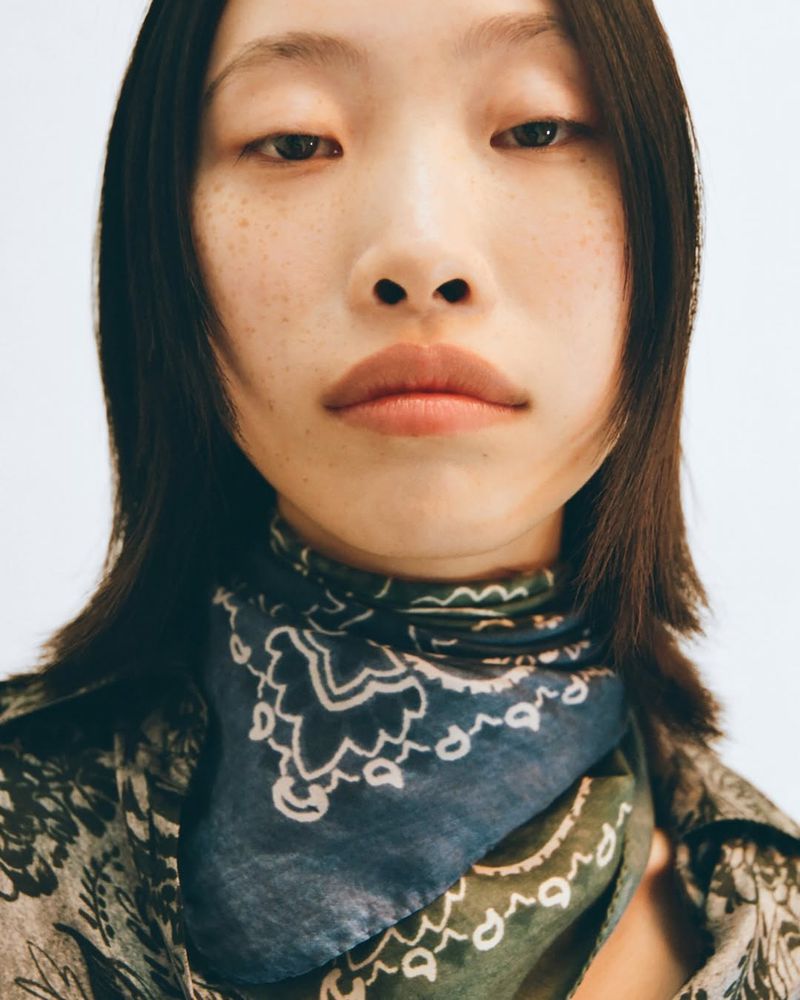 Close-up portrait of a woman with freckles wearing a patterned shirt and bandana, gazing directly at the camera against a bright background.