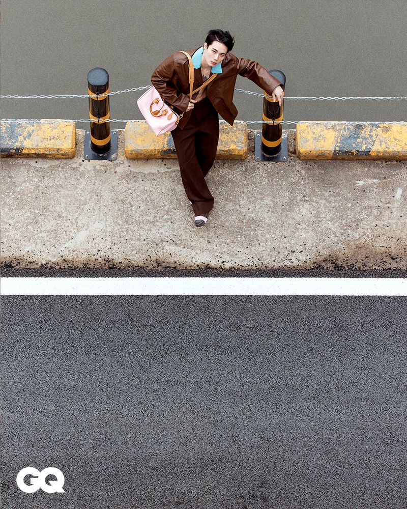 Stylish young man in brown leather jacket and pants poses on a concrete structure next to a road with a pink purse.