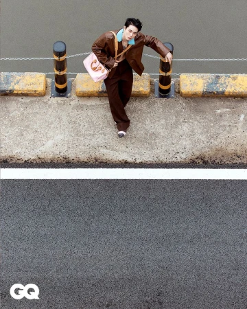 Stylish young man in brown leather jacket and pants poses on a concrete structure next to a road with a pink purse.