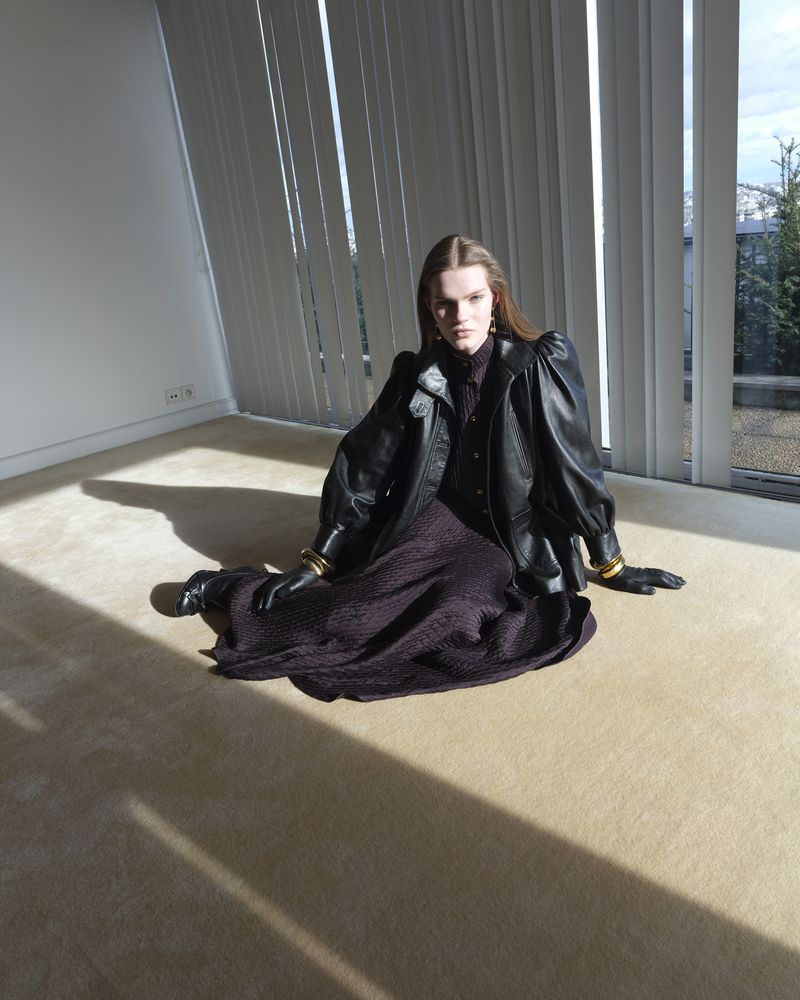 A fashionable woman in a black leather jacket and a dark dress sits on a light carpet in a minimalist room with sunlight streaming in.