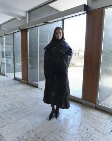 Model standing in a long dark coat inside a modern building interior with large glass doors and marble flooring