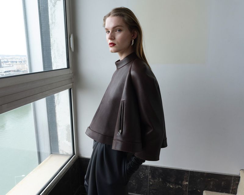 Indoor shot of a fashionable adult female model in a brown leather jacket and gray pants posing near a window with an urban view.
