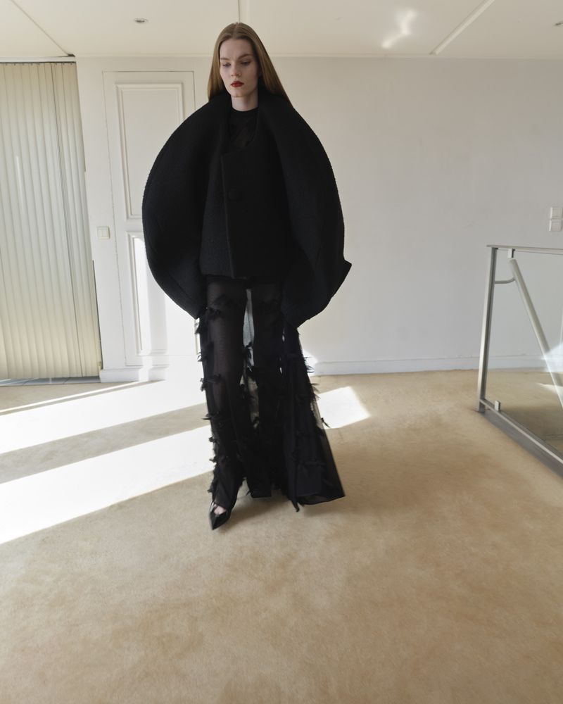 A fashionable adult woman with long red hair poses in a large black avant-garde coat and matching sheer, fringed pants in a bright, modern interior.