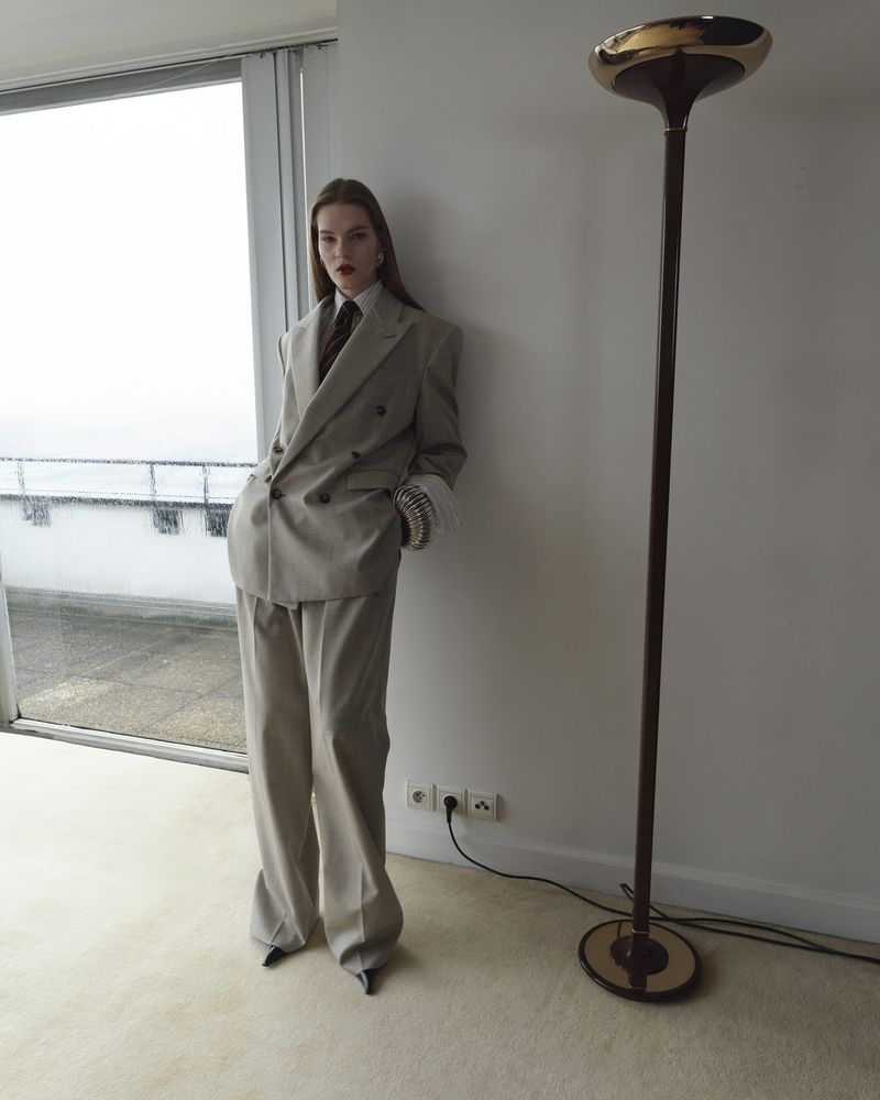 A young adult female model poses indoors, wearing a gray pantsuit with a tie. A modern floor lamp is visible against a neutral background.