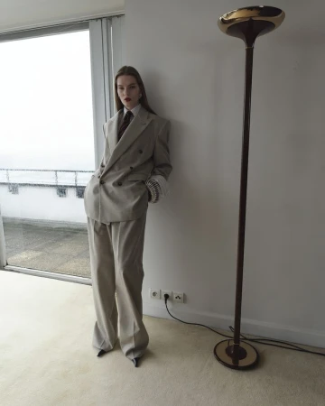 A young adult female model poses indoors, wearing a gray pantsuit with a tie. A modern floor lamp is visible against a neutral background.