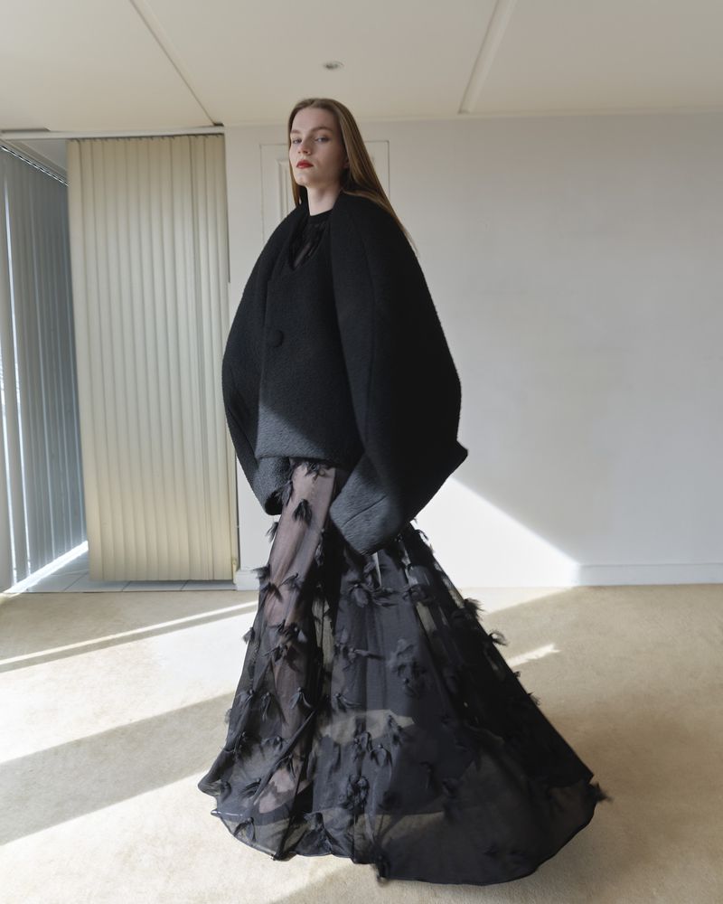 A full shot of an adult female fashion model posing indoors, wearing a black cape-like jacket and long black tulle skirt.