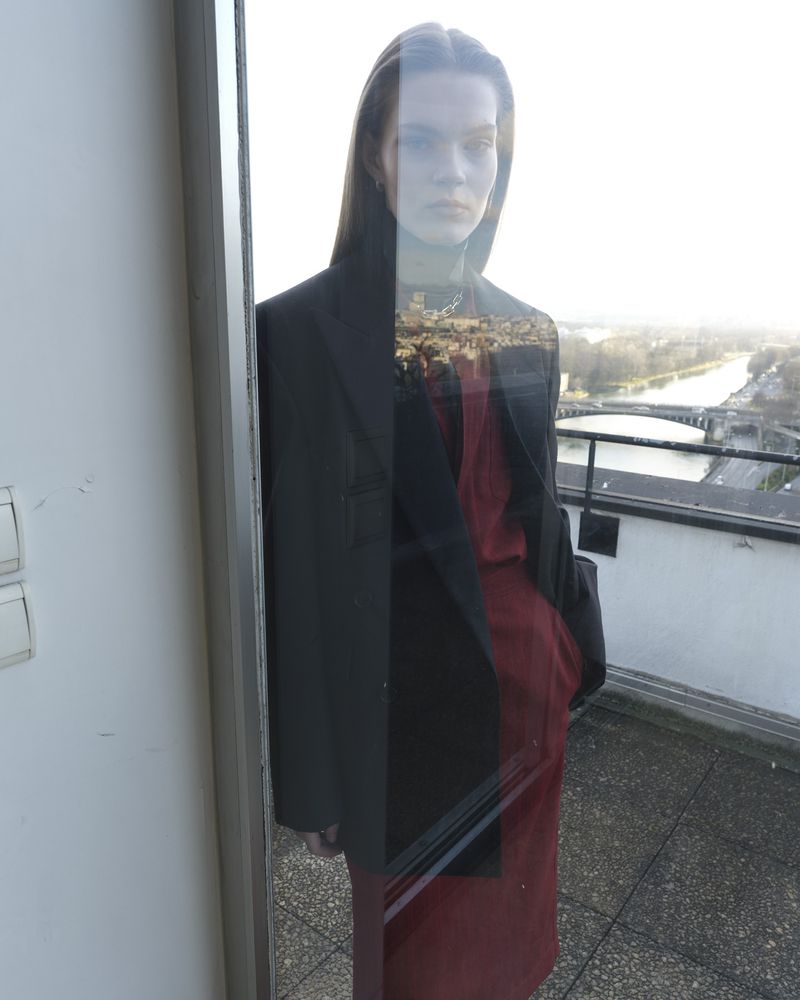 Model standing on a balcony by a window, wearing a dark blazer over a red outfit with a distant cityscape in the background.