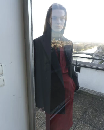 Model standing on a balcony by a window, wearing a dark blazer over a red outfit with a distant cityscape in the background.