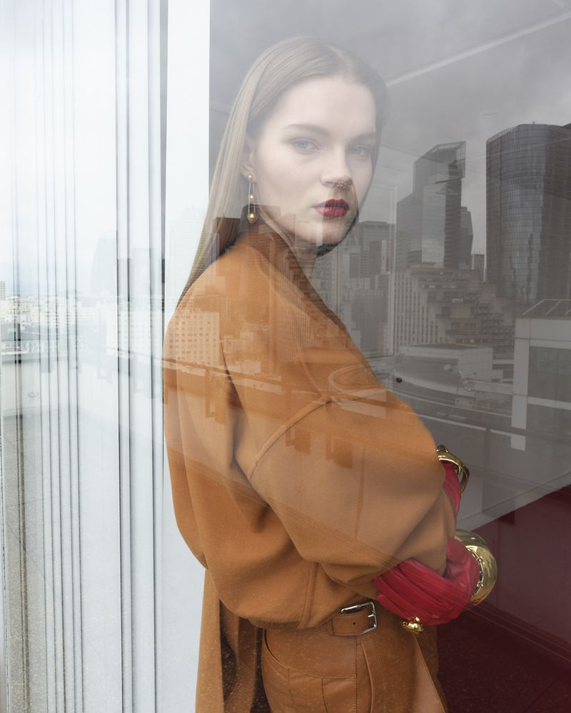 A woman in a camel coat and red gloves stands in front of a window with a cityscape reflection, creating a blurred urban background.