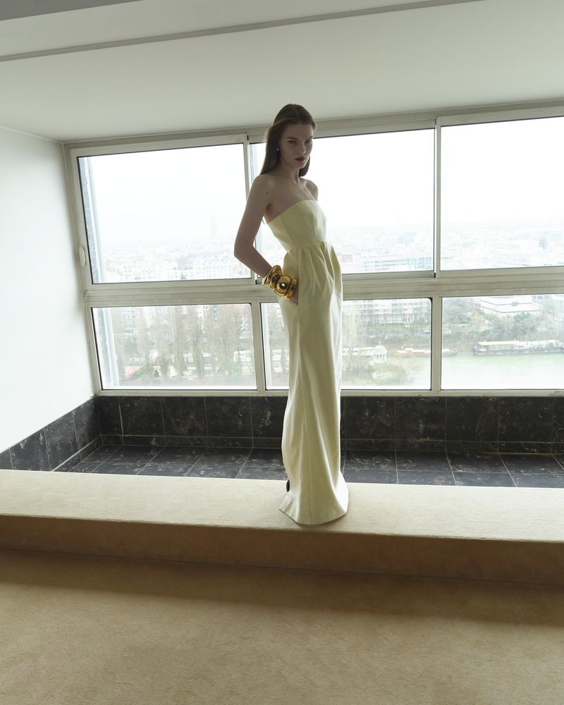 Indoor fashion shot of a young woman in a long strapless yellow dress with gold bracelets, posing near a large window with a cityscape view.