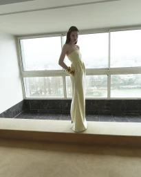 Model standing indoors wearing a long pale yellow strapless gown against large horizontal windows.