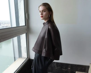 Model wearing a dark brown leather high-neck cape and trousers standing near a large window for T Magazine China
