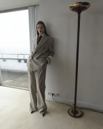 Model leaning against a white wall wearing an oversized beige pantsuit and tie, next to a tall floor lamp.