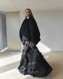Model wearing a structured black coat and sheer feathered gown inside a minimalist interior