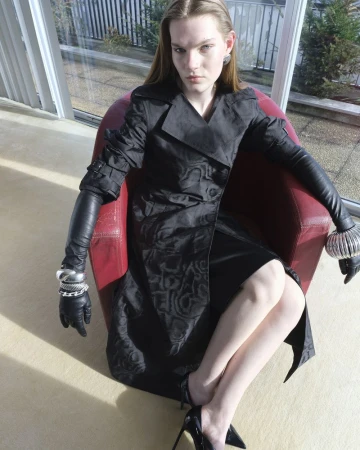 Model seated in a red armchair wearing a black patterned coat, long black leather gloves, and silver bracelets against a bright window.