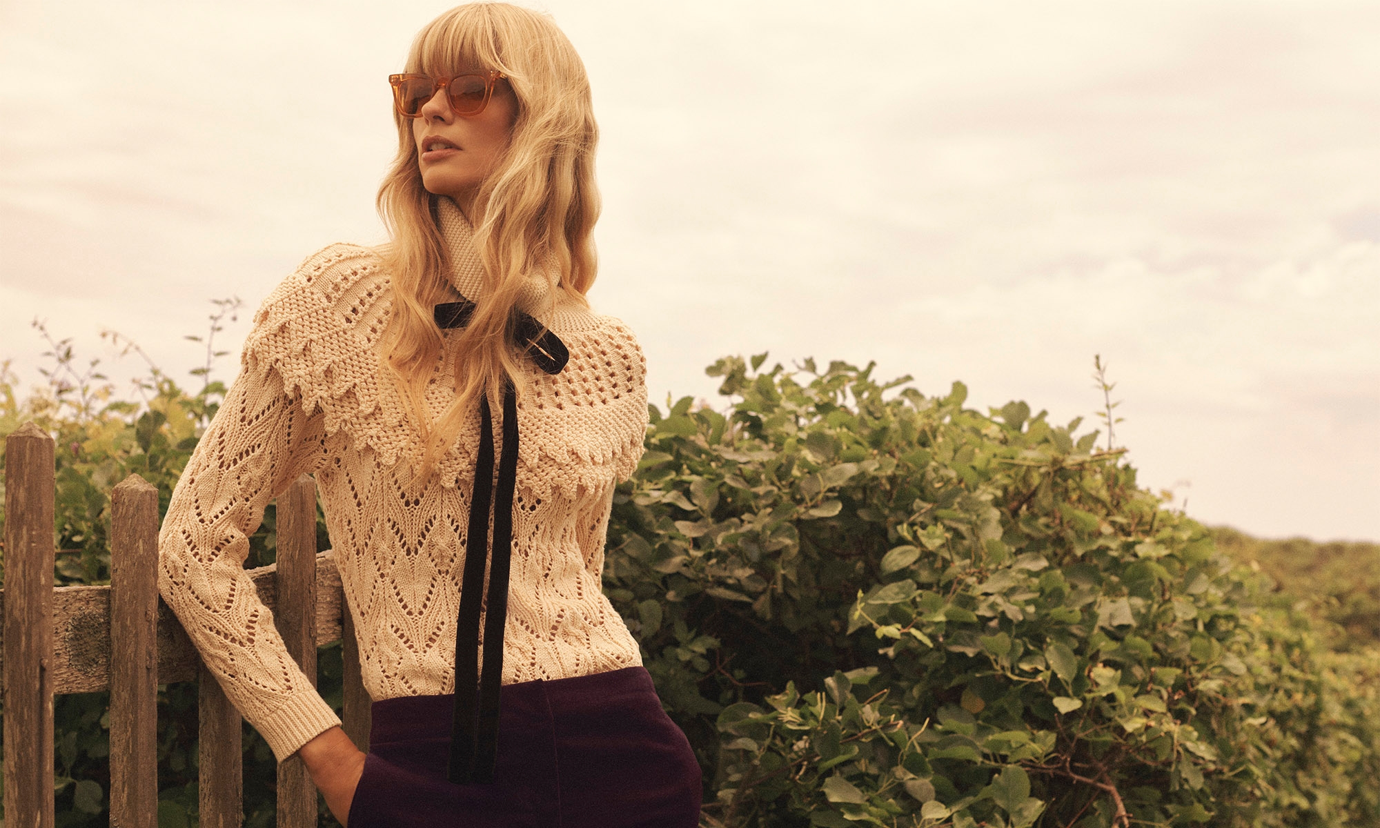A fair-skinned woman poses outdoors wearing an eyelet sweater with a black bow, maroon trousers, and tinted sunglasses; medium shot.