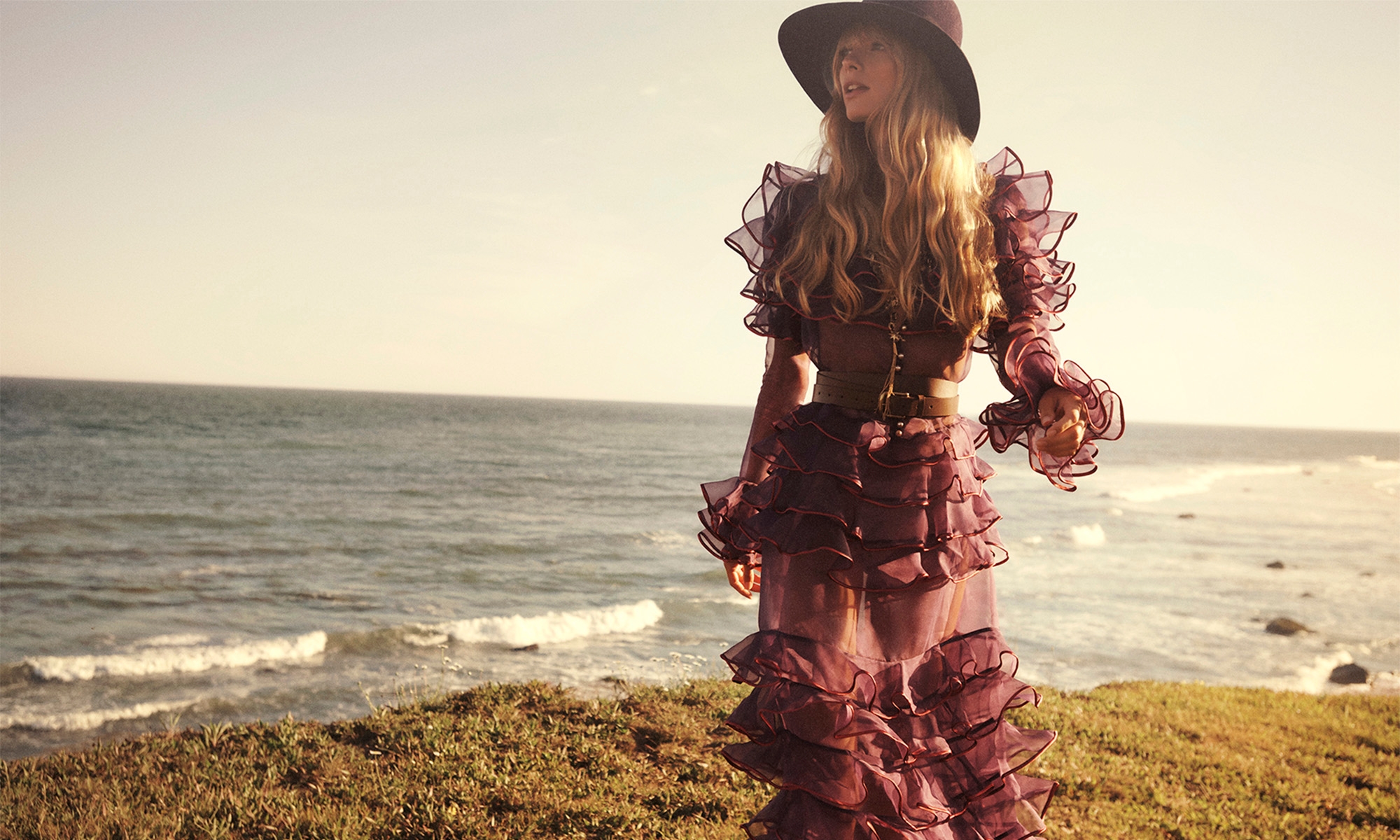 Woman wearing a sheer purple ruffled dress and wide-brimmed hat standing on a grassy coastal cliff overlooking the ocean.