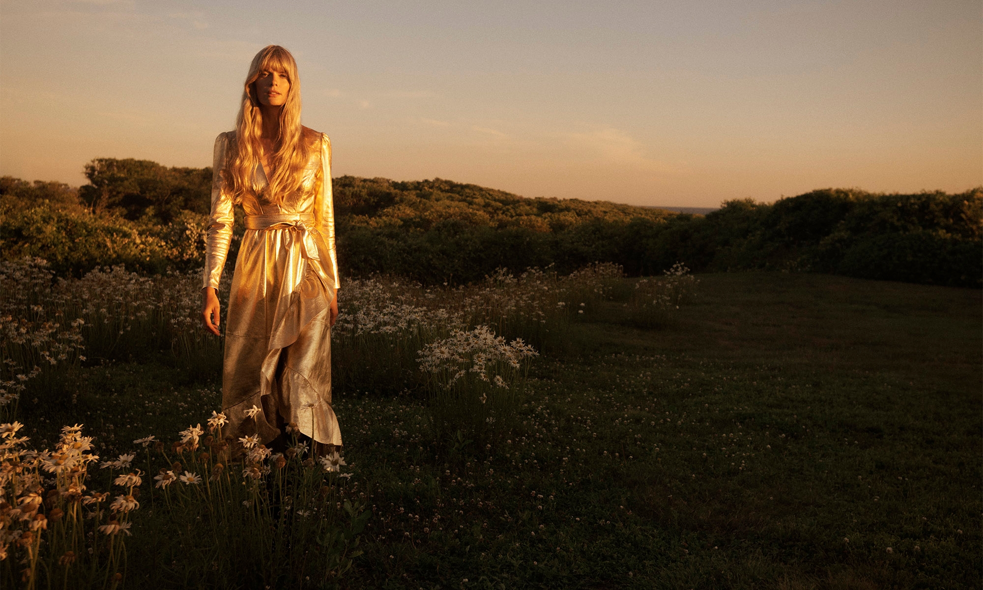 Model wearing a gold metallic wrap dress standing in a wildflower field at sunset for Zimmermann Fall RTW.