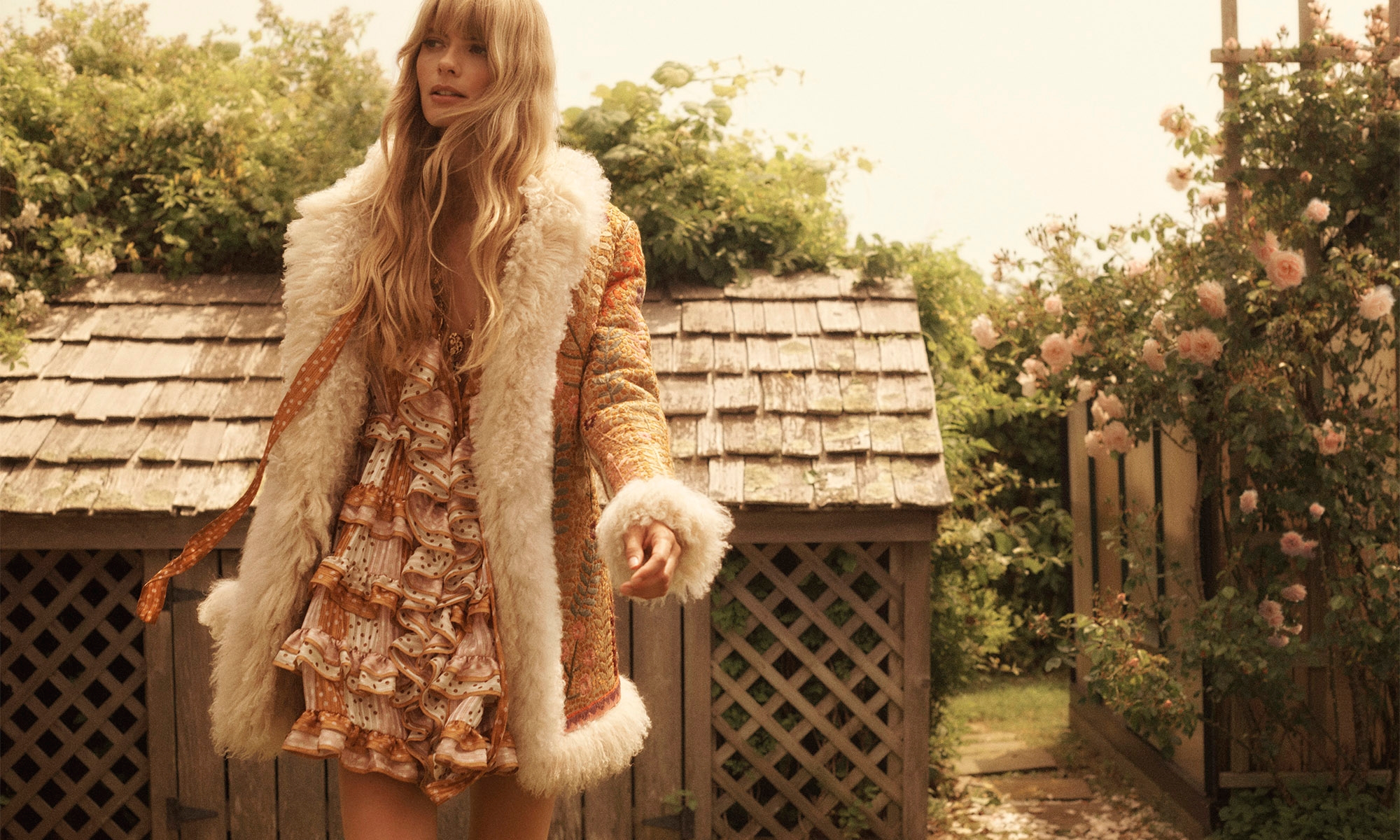 A blonde woman in a vintage-style dress and fur-collared coat stands outdoors surrounded by greenery and flowers on a sunny day.