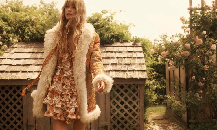 A blonde woman in a vintage-style dress and fur-collared coat stands outdoors surrounded by greenery and flowers on a sunny day.