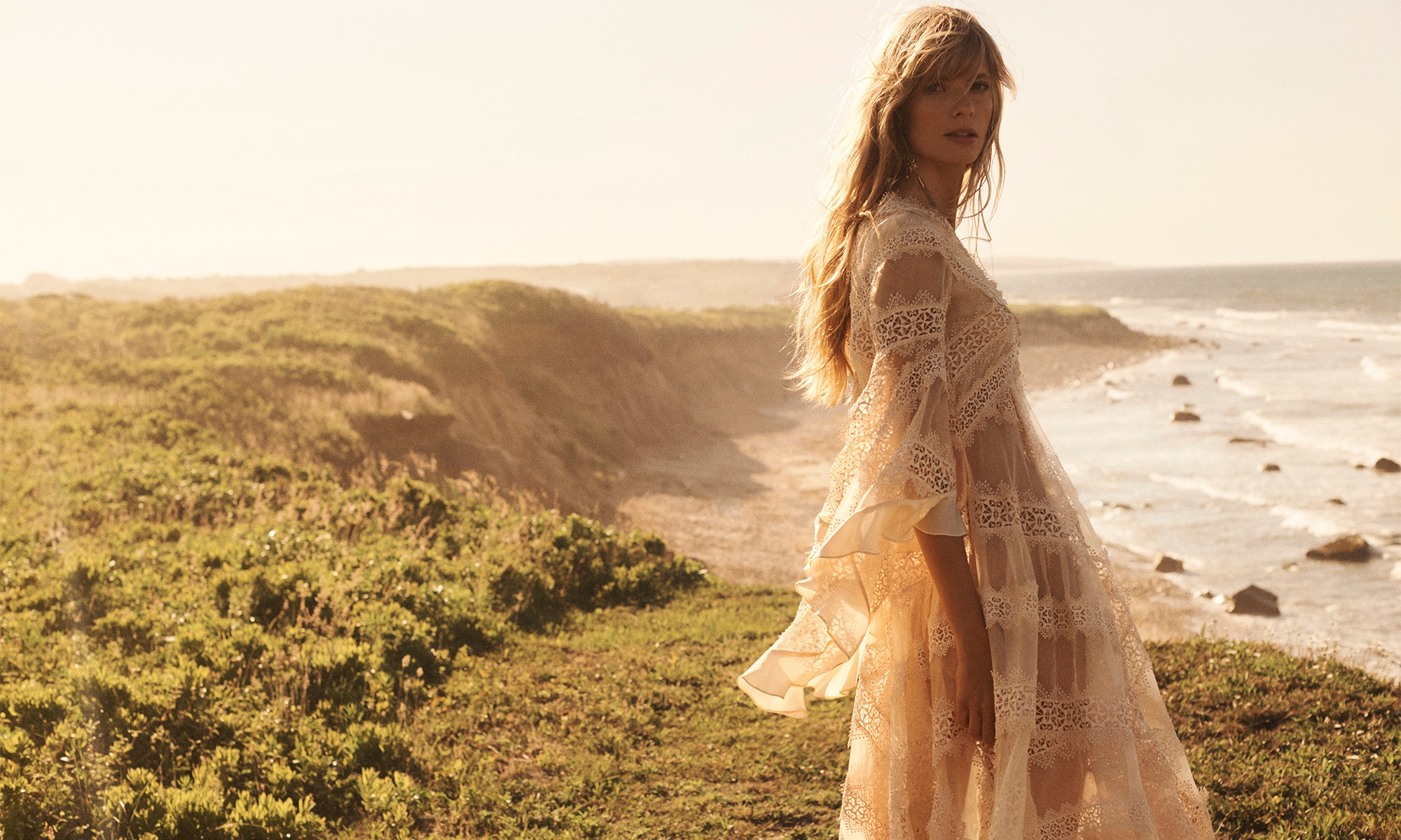 A woman in a lace dress stands on a grassy cliff overlooking the ocean during daytime. Sunlight creates a warm, dreamy atmosphere.