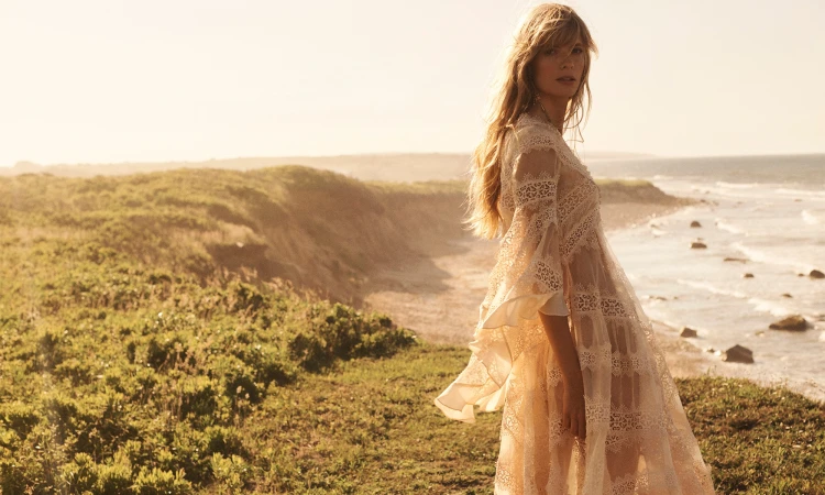 A woman in a lace dress stands on a grassy cliff overlooking the ocean during daytime. Sunlight creates a warm, dreamy atmosphere.