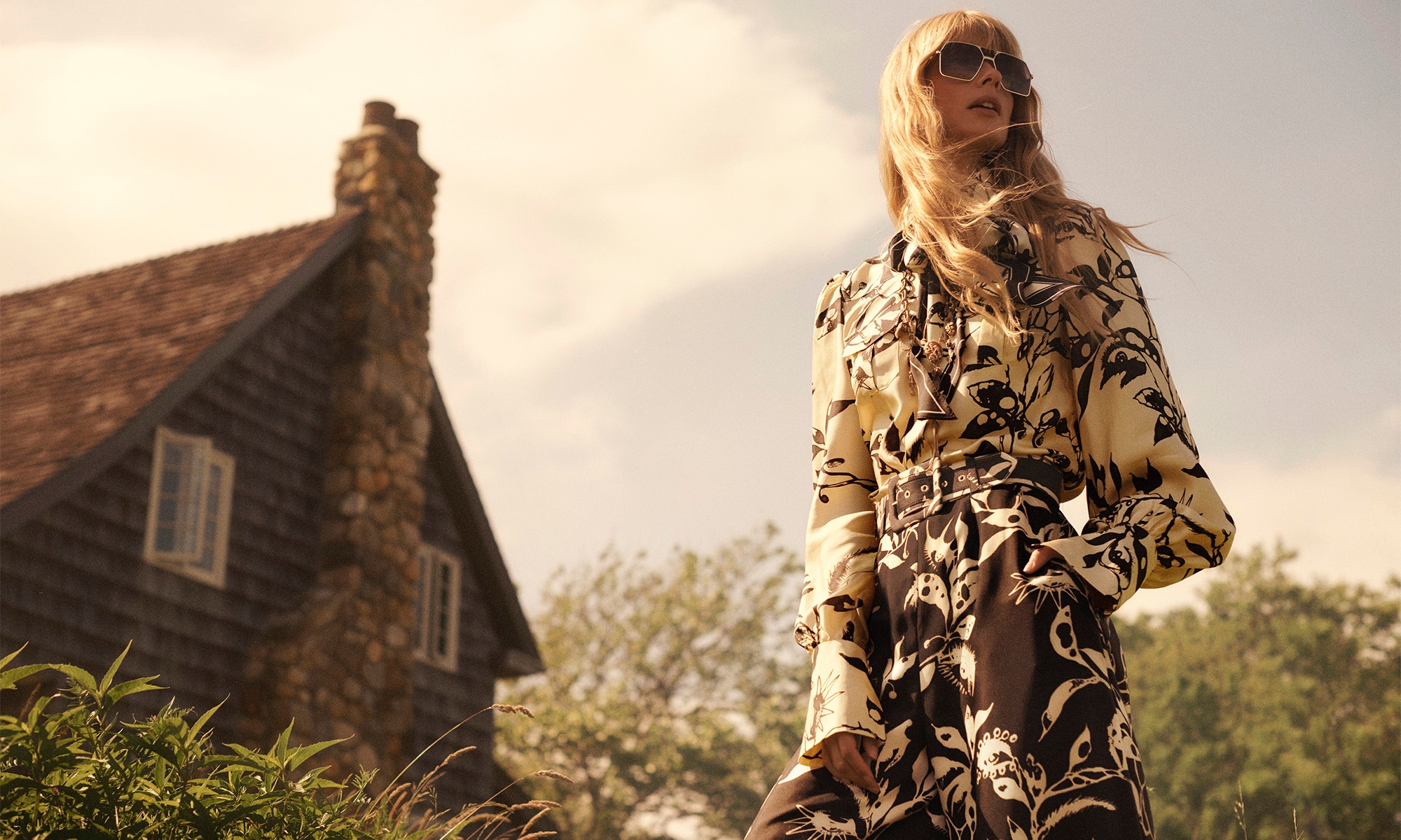 A blonde woman in a patterned pantsuit and sunglasses stands outdoors near a rustic wood house on a sunny day, fashion shoot.