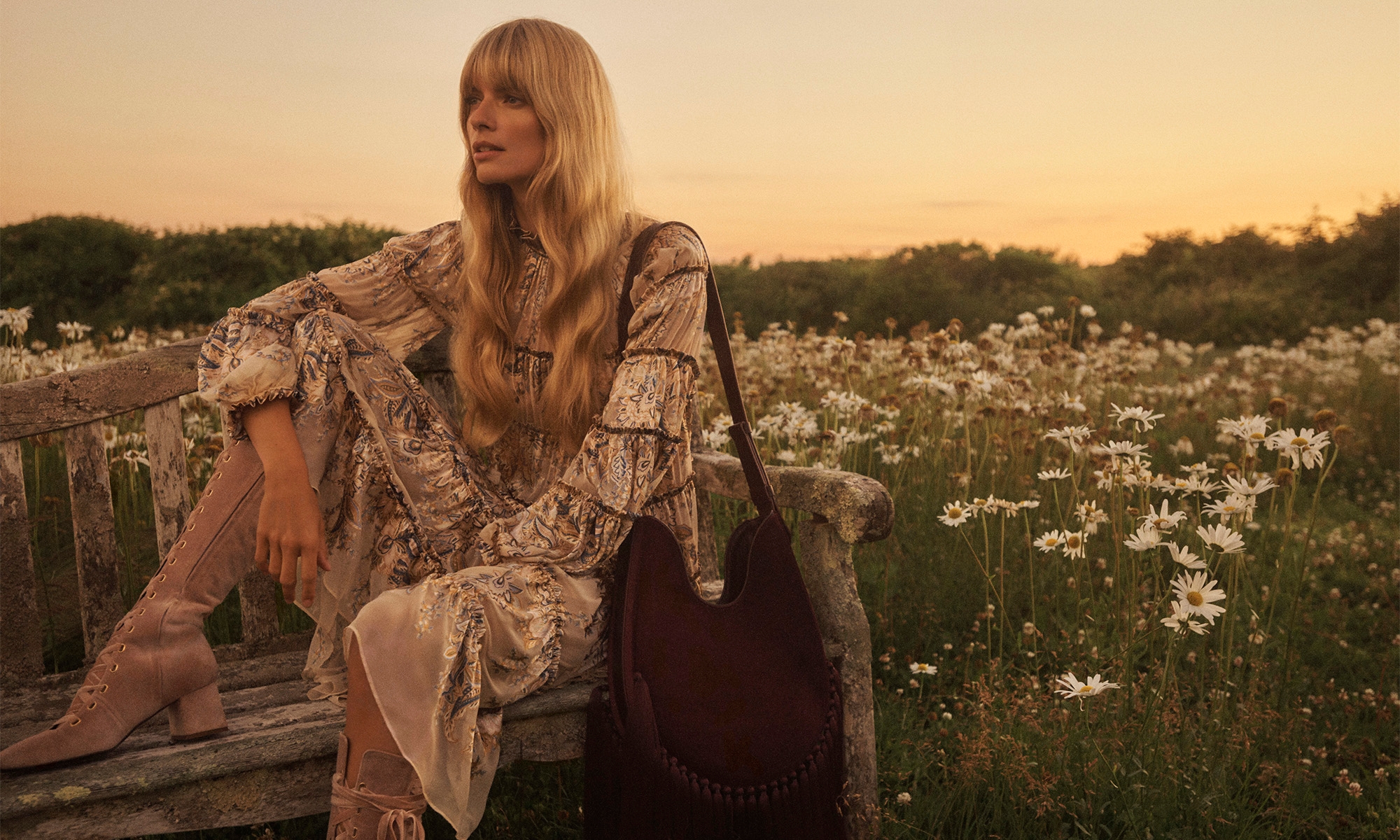 Julia Stegner seated on a weathered wooden bench in a field of daisies wearing a patterned dress during sunset