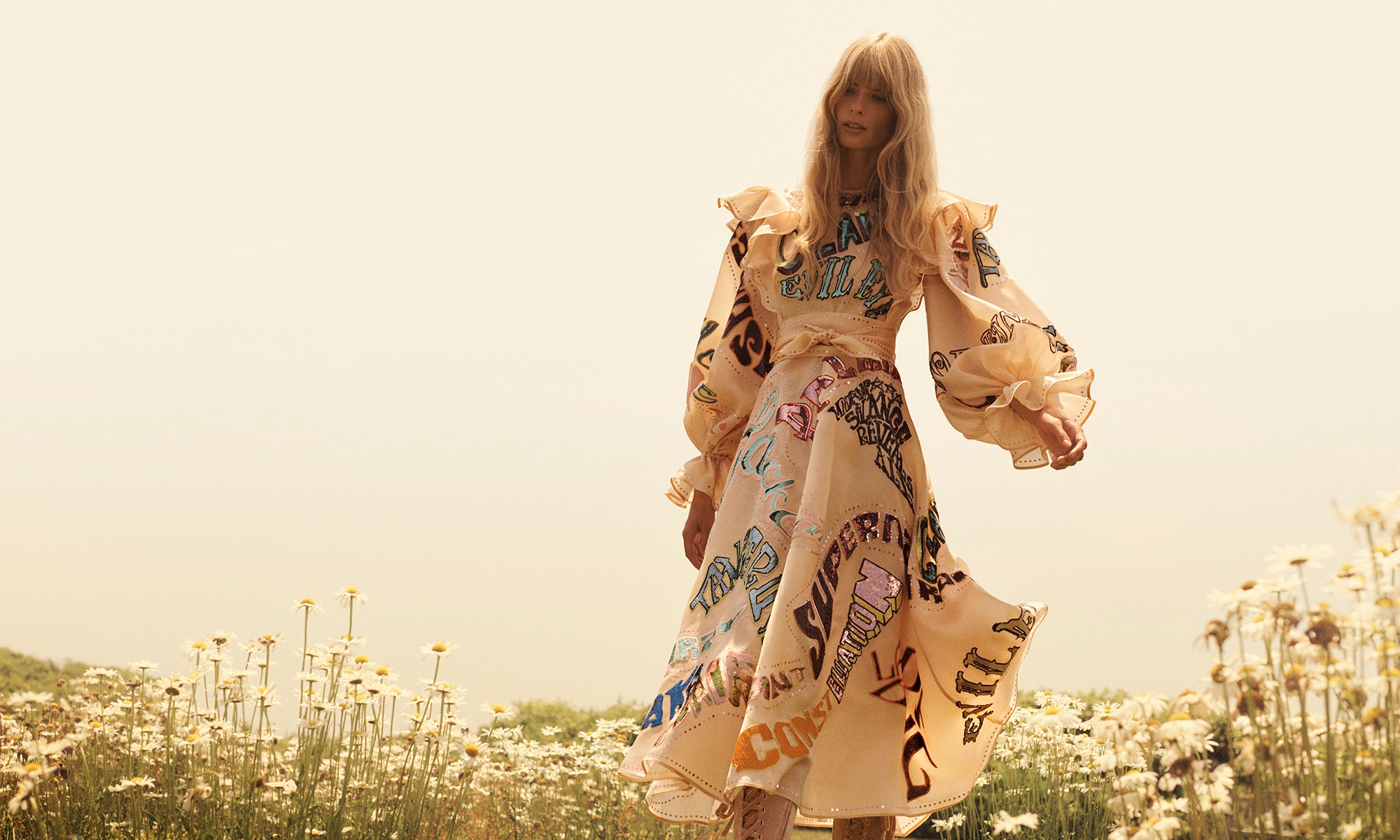 Adult female model in patterned ruffle dress poses in outdoor field of daisies during daytime with fair skin and blonde hair.