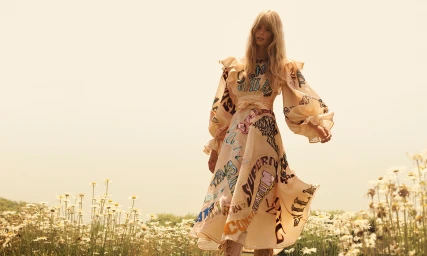Model Julia Stegner wearing a cream ruffled dress standing in a meadow filled with white flowers under a warm sky.