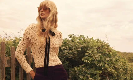Model in cream knitted sweater with black ribbon tie and sunglasses leaning on wooden fence by foliage