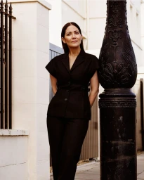 Mishal Husain posing outdoors in a black tailored jacket and trousers leaning against a decorative lamppost for British Vogue