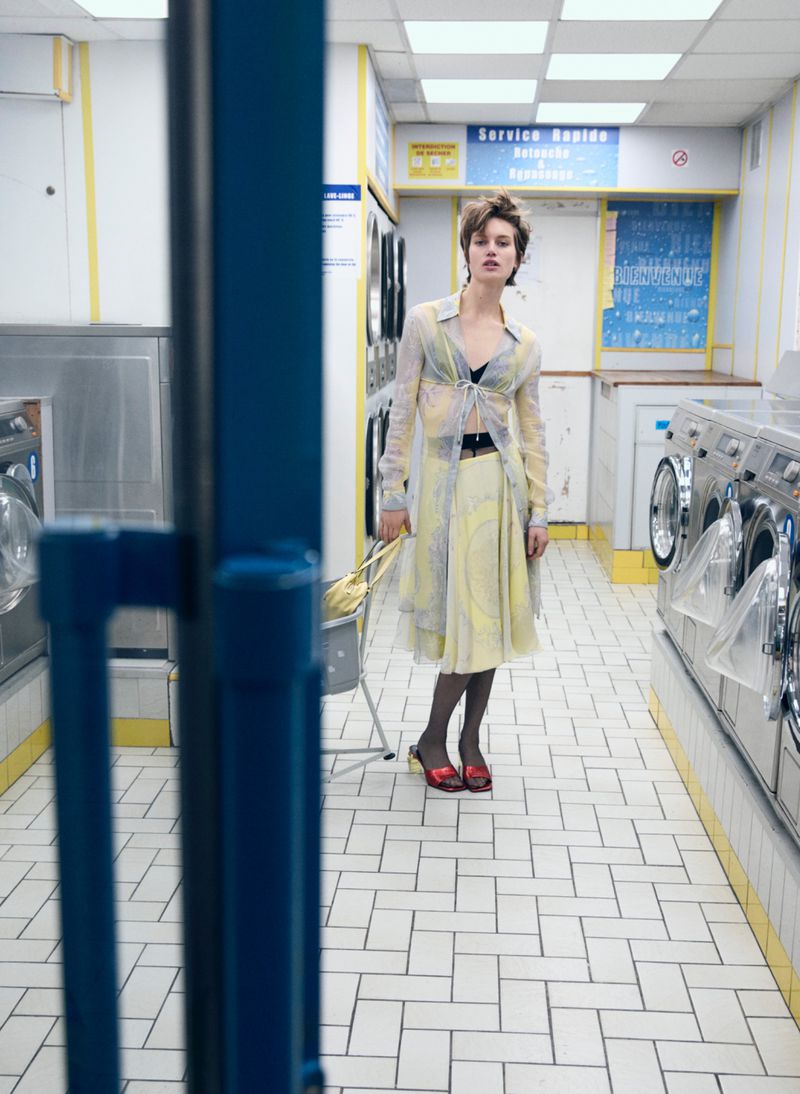 A young woman stands in a brightly lit laundromat wearing a semi-sheer top and skirt, dark tights, and red heels.
